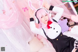 A woman with pink hair sitting on a bed wearing headphones.